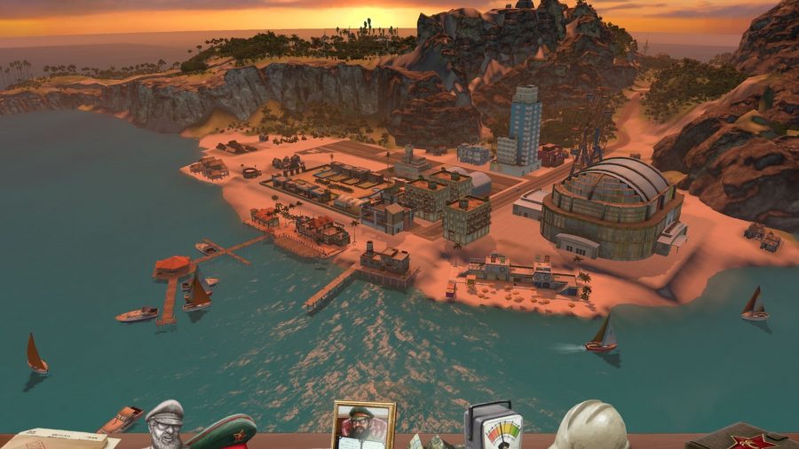 A screenshot from Tropico showing a tropical island, with various buildings by the sea, and trees in the distance.