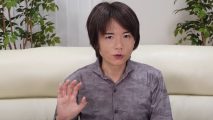 A picture of Masahiro Sakurai with on hand raised, wearing a purple button-up shirt. He has long-ish black hair parted in the middle.
