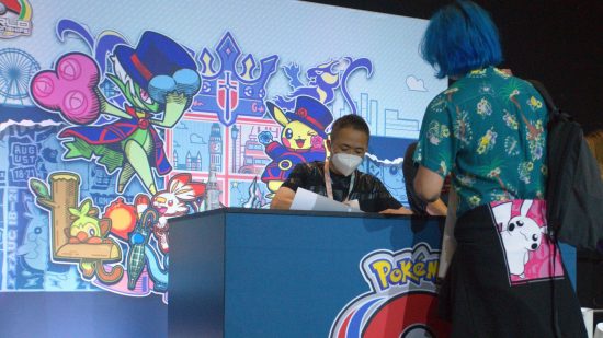Pokemon Worlds 2022: Junichi Masuda sits at a desk wearing a mask, signing posters for an eager fan