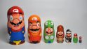 Various Mario matryoshka dolls lined up. From right to left: a large blue dungaree-wearing Mario, a smaller one with an orange hat, a smaller, yoshi-esque Mario, an even smaller brown Mario, and a tiny black Mario.