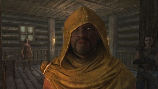 Skyrim marriage: a man with bright eyes looks out from beneath a hooded yellow robe in a wooden walled room.
