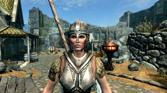 Skyrim's Lydia stood in a town surrounded by walls, with fire torches, leaves on the ground, and other medieval-esque elements. She is a woman in silver armour, with a silver helmet on her head and axe on her back.
