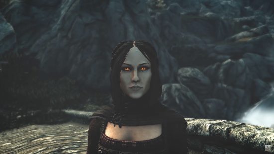 A shot of Skyrim's Serana, a woman with glowing red eyes, white face, and red robe and hood, stood on a stoney surface.