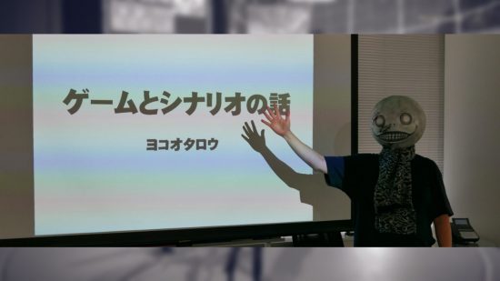 Yoko Taro, wearing the Emil mask from Nier Automata, a spherical head with a grin and wide eyes -- he has his hand raised towards a whiteboard with Japanese writing on it, and a black and white scarf over a black jumper.