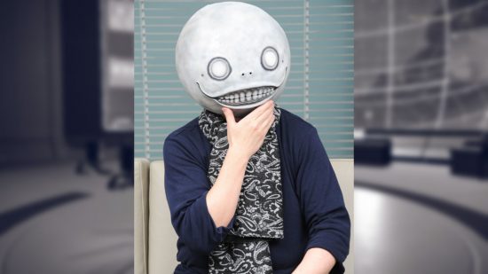 Yoko Taro, wearing the Emil mask from Nier Automata, a spherical head with a grin and wide eyes -- he has his hand to its chin like he's thinking, and a black and white scarf over a black jumper.
