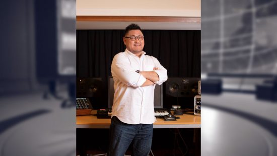 Keiichi Okabe, composer on Nier Automata, with his arms crossed in a white shirt and jeans, standing in front of a wooden desk with music equipment on it.