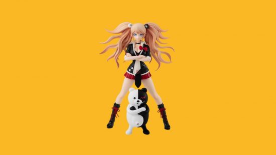 A Danganronpa figure on a yellow background. It's a woman with wild ginger hair in a red skirt and school blazer, knee high boots, and a teddy bear below. Both are stood legs apart and arms folded.