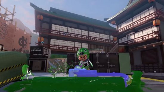 Splatoon 3 song titles: a Splatoon character in a green scrum hat (like, for rugby), holds a massive paint roller and splats green ink on the ground in a large, closed in space with tall wooden buildings behind.