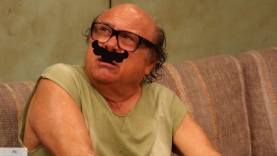 Danny DeVito in a vest looking sweaty with glasses and bald top of head on a sofa, with a black cartoon moustache superimposed over the top.