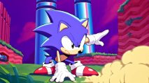 Sonic Origins Plus release date trailer screenshot showing Sonic, a cartoon blue hedgehog in red shoes and white gloves, skidding across some grass with tall blue pipes behind him in a red and purple sky.