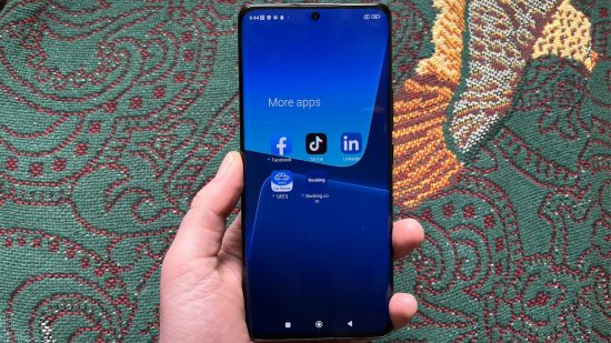Xiaomi 13 Pro review - the Xiaomi 13 Pro, a large smartphone with a black ceramic back, aluminium sides, and glass front, held in a hand face up, showing the home screen, blue background, and various apps, with a green and red throw behind it.