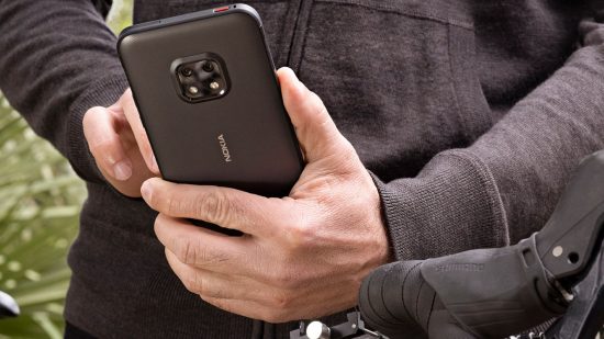 Jeden z najlepszych wytrzymałych telefonów, czarna Nokia Xr20 w męskiej dłoni.. Zwykły z aparatem na górze pośrodku.  mężczyzna ma białe ręce i ma na sobie czarną bluzę z kapturem przy rowerze.