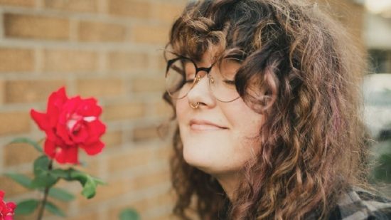 Whitethorn Games chief accessibility officer: Britt Dye, a white woman with brown curly hair and glasses, sniffing a red flower.