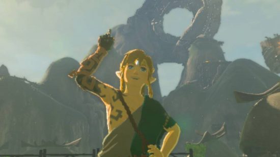 Zelda Tears of the Kingdom paraglider - Link, a blonde boy in a circlet and green and grey tunic with strange markings on his right arm which is raised aloft with one finger raised in a jovial pose.