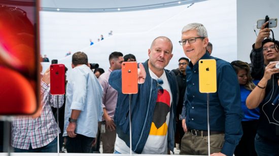 Apple ChatGPT -- Jony Ive, a bald man in a blue jacket and whit shirt with abstract design on the front, pointing at an orange iPhone on a stand in a crowded room, standing next to Tim Cook, a man with grey hair and glasses in a dark shirt, stood in front of a yellow iPhone on a stand.