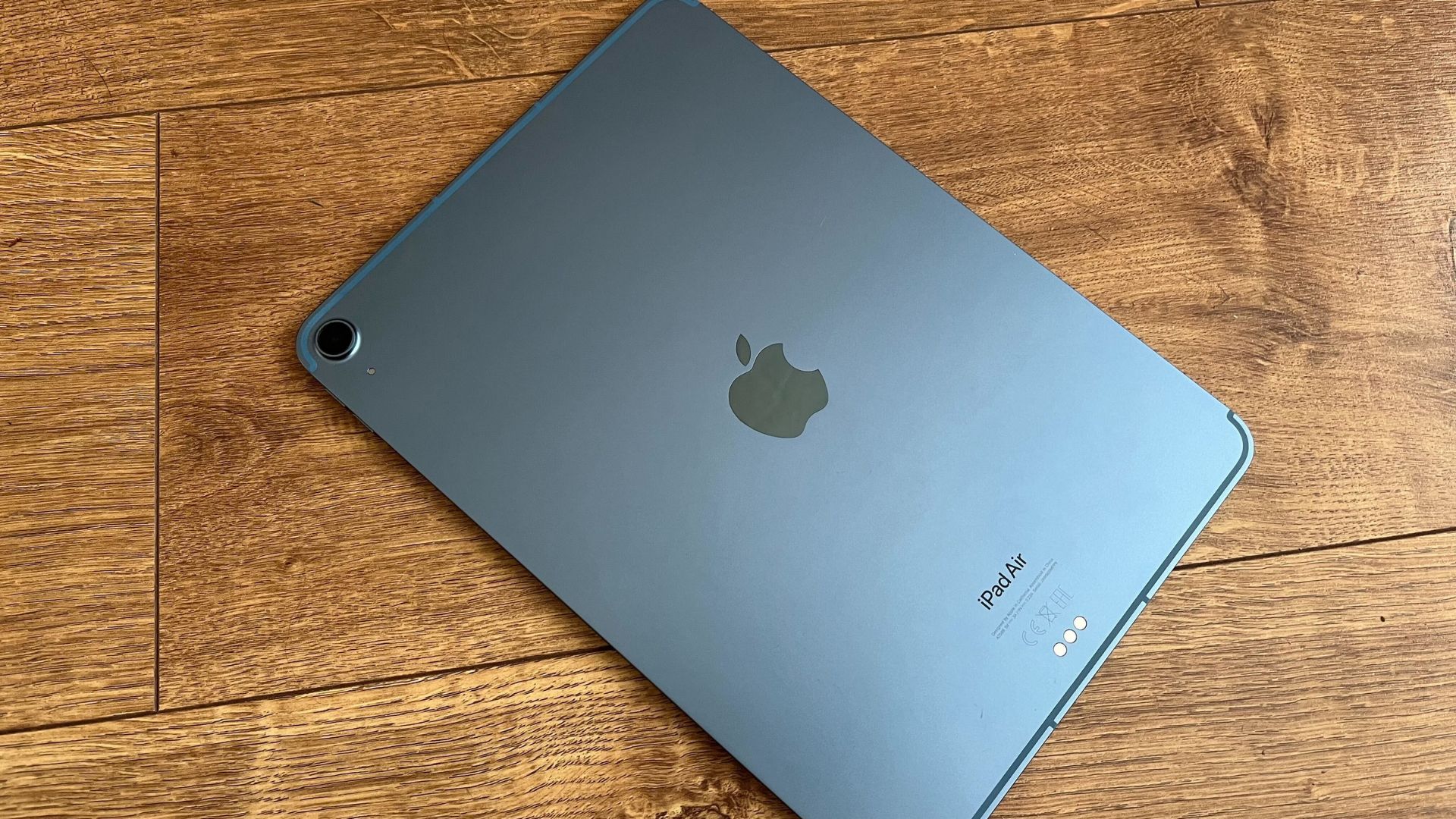 One of the best iPads, the iPad Air, in black lay on its screen on a wood surface. We see its back, showing the Apple logo in the centre and small single camera ring in the top right corner.