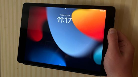 iPad 9 Gen review shot, showing the iPad screen with a black red blue and orange abstract background and time showing, held in landscape, with a hand holding it on the right hand side.
