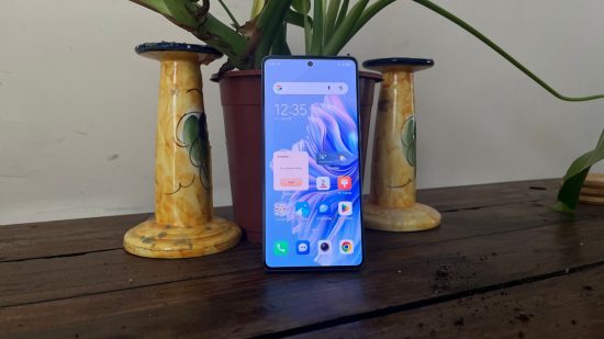 Tecno Camon 20 Premier review - the phone leant on a plant between two candlesticks. The phone is unlocked showing the home screen apps and blue abstract background.