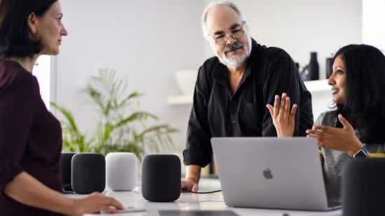 Apple AI header showing three people around a table with three HomePod speakers and a MacBook, looking like they're all having an important meeting.