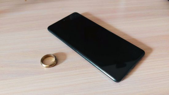 Samsung ring header showing a gold ring and a large phone with the screen off lay flat on a faux-wood desk.