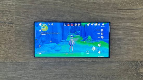 Samsung Galaxy S23 Ultra review header showing the phone's screen with Genshin Impact playing, phone lay flat on a wooden surface.