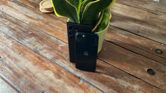iPhone vs. Samsung: which is better for gaming? header image showing two phones, the iPhone 13 and the Samsung S23 Ultra. They're both black. Leaning against a plant, the iPhone is in front of the Samsung, both showing the back.