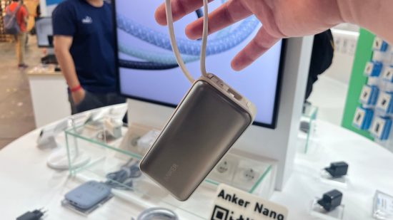 Anker USB-C power bank dangling from its cable in front of a screen and a table. It is white and silver and square.