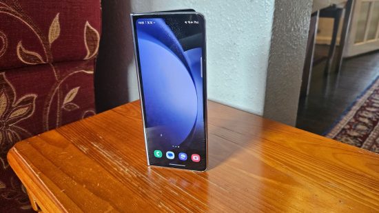Samsung Galaxy Z Fold5 review header showing the phone in blue half folded standing upright on a wooden table with the screen showing a background.