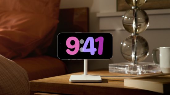 What is iOS: A photograph of someone's bedside table featuring a landscape iPhone on a MagSafe stand with a fullscreen landscape digital clock display in various shades of purple on a black background