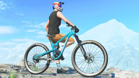 A person leaning on their bike in shorts, a vest, and a redbull helmet in front of a mountain