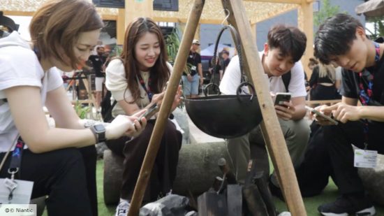 Monster Hunter Now interview - a photograph from Niantic showing a group of people sitting around a replica camp fire from Monster Hunter playing the game on their phones and smiling