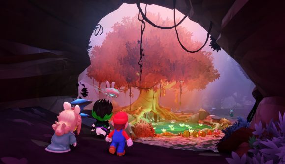 Screenshot from Mario + Rabbids Sparks of Hope showing Mario and a Rabbit dressed as peach walking through a cave (their backs to us) towards a red tree in bloom. Vines hang from the cave ceiling, and mossy stuff covers the base of the tree.