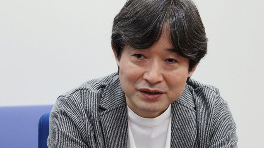 Tetsuya Takahashi, creator of Xenoblade Chronicles, in a white shirt and grey jacket. In is a middle-aged Japanese man with dark brown (maybe black) hair.